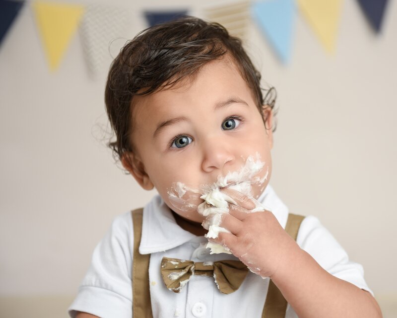 A baby boy in a white shirt and suspenders, with green eyes and frosting on his face, enjoying his cake smash session in a pastel-themed setup with bunting.