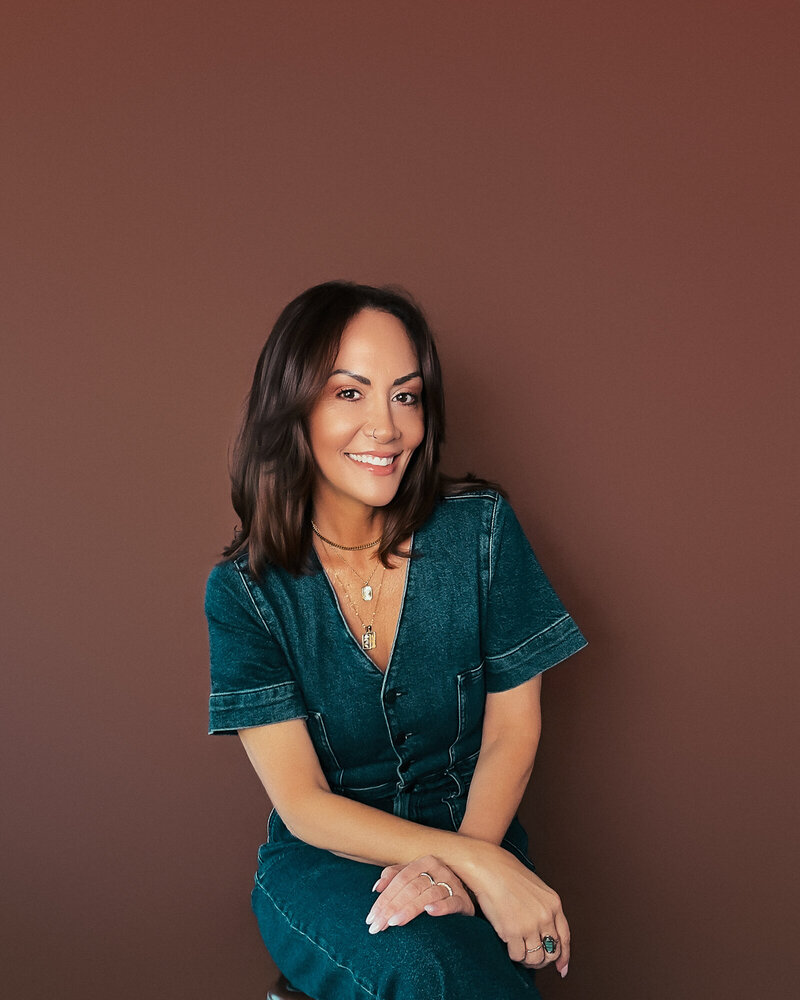 brunette woman on a brown background wearing a jean material jumpsuit and smiling at camera