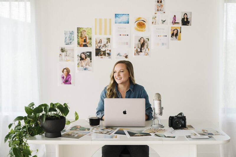Krista Marie, sitting at a desk with photos taped to the wall, working on her laptop, smiling and looking away from the camera