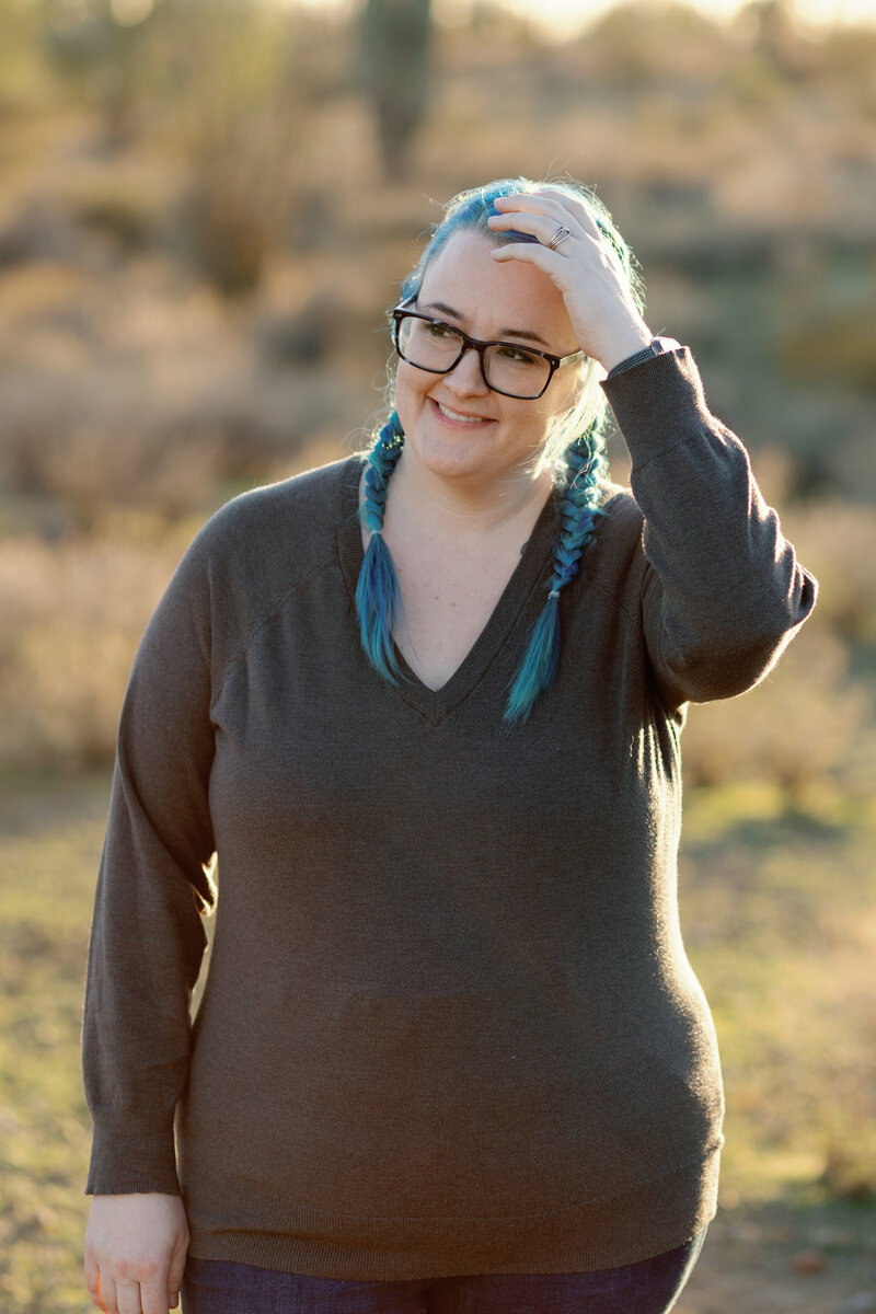 Woman with blue hair in french braids with rectangular dark blue glasses and a grey sweater looking off in the distance fixing her bangs with her hand