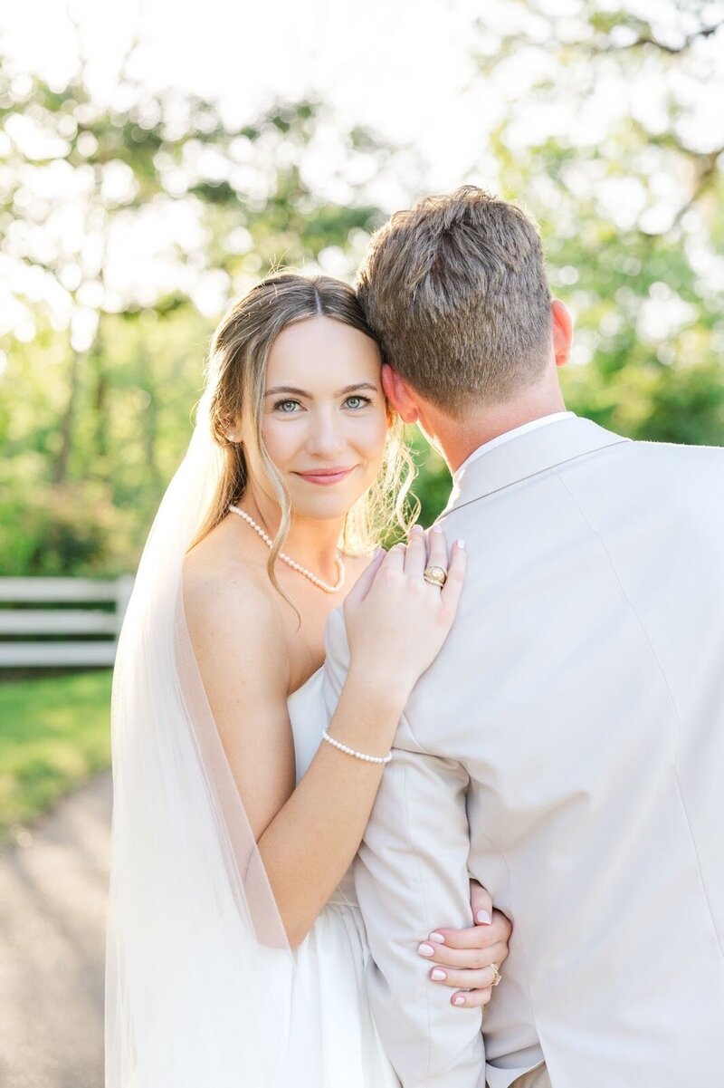 Bride wears a San Antonio wedding dress and veil while looking over her groom's shoulder down at the camera smiling. The dress includes an off the shoulder look with pearls.