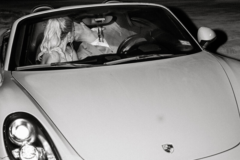 Black and white image of bride and groom kissing in a convertible leaving their wedding