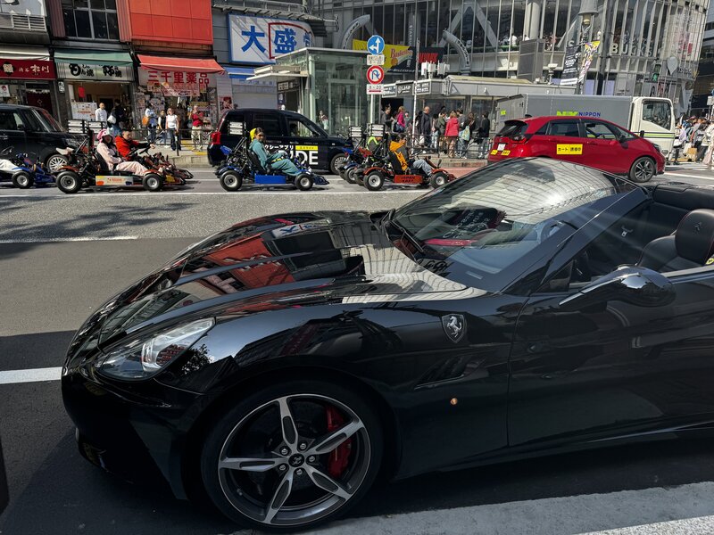 Black Ferrari parked on a busy Tokyo street with locals, scooters, and colorful storefronts in the background, capturing city energy and luxury vibes.