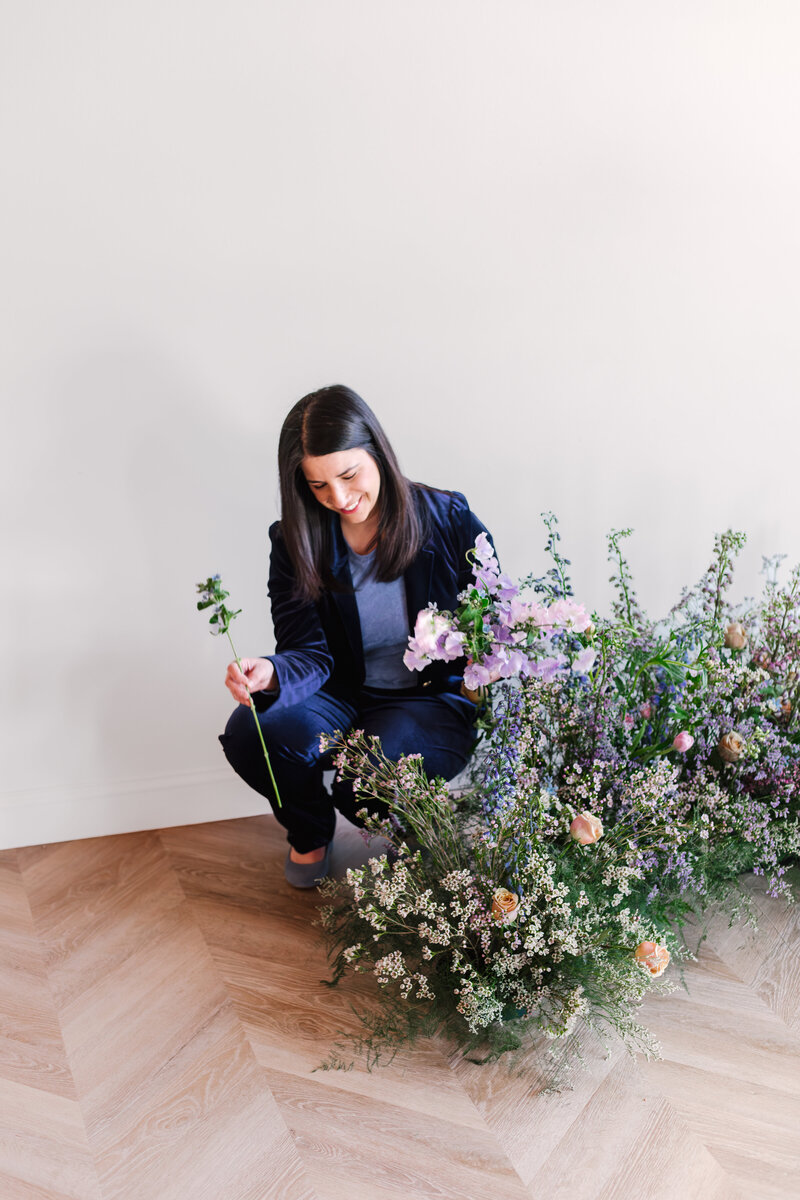 Florist next to floral arrangement, arranging blooms with a smile against a plain backdrop.