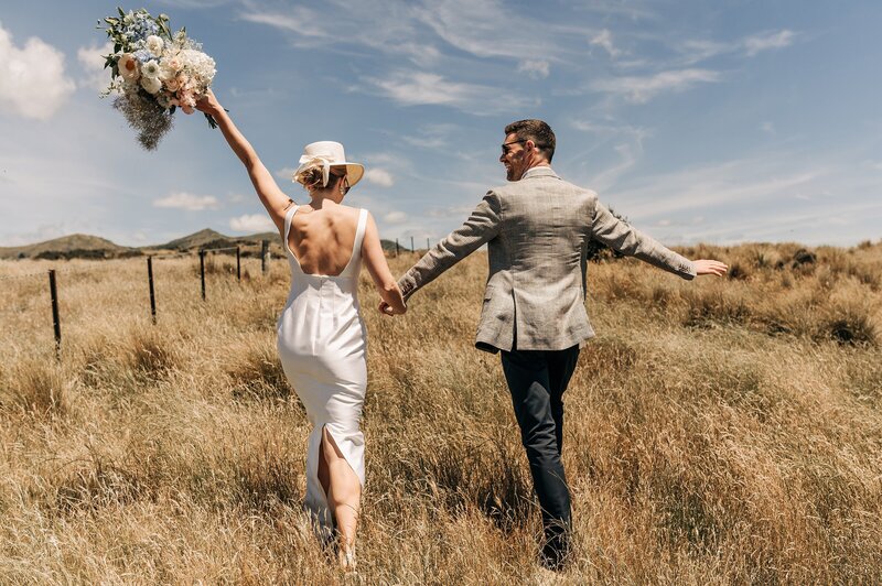 a bride in emilia wickstead dress and cream hat walk through golden grass on a sunny day in blenheim