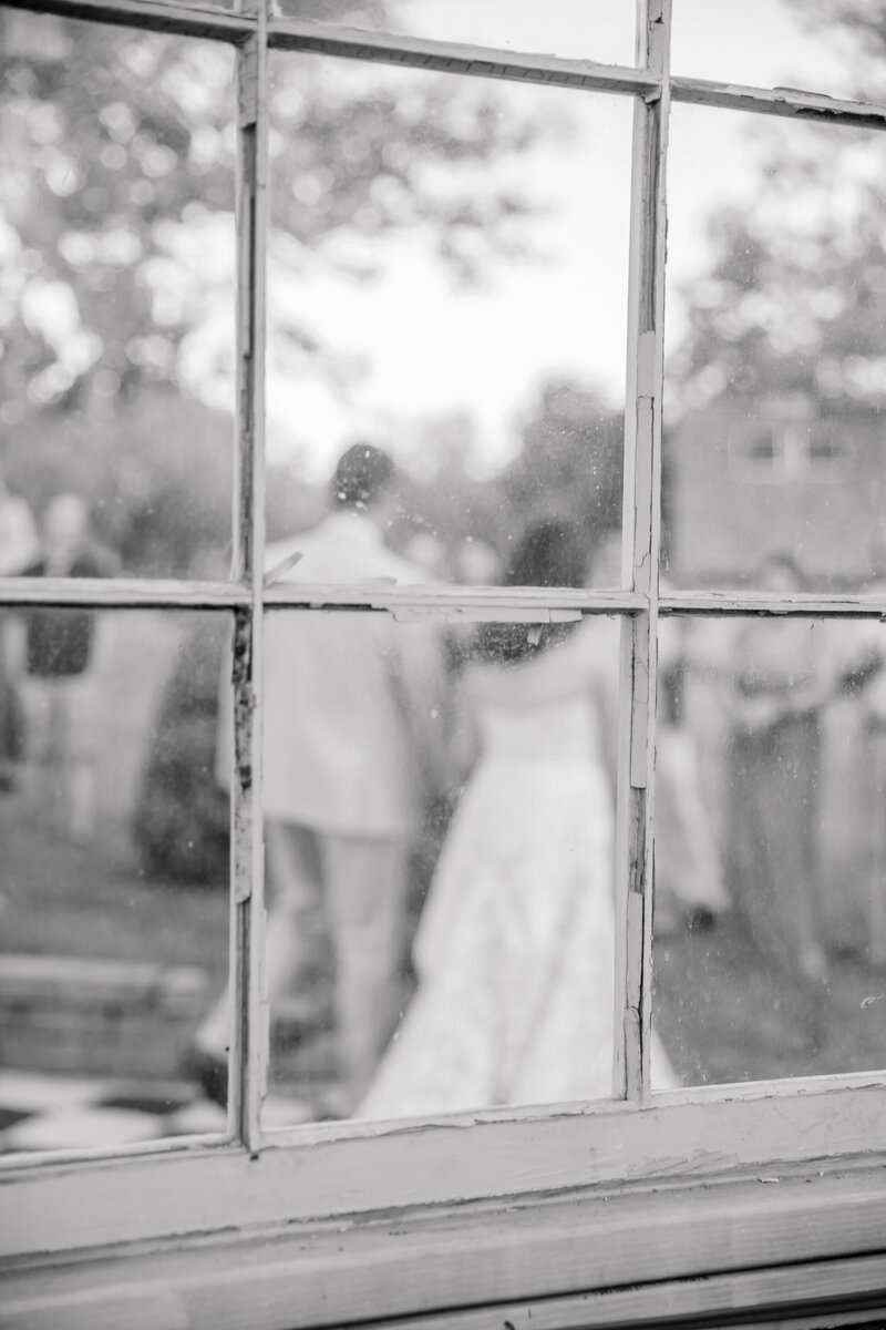 Black and white image through the window of the greenhouse at a Three Oaks Manor wedding. The bride and groom exit the dance floor hand in hand after their first dance.