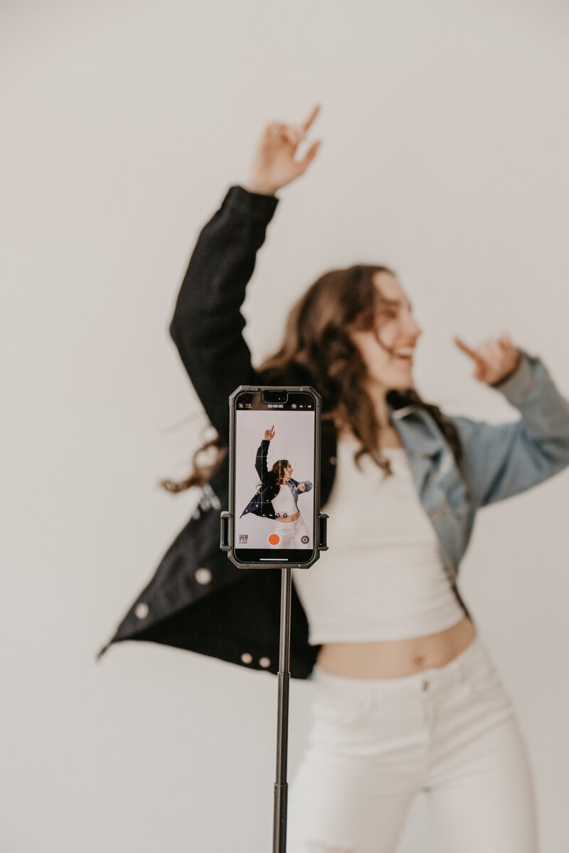 A UGC content creator dancing in front of her tripod and phone, which is recording her dance. She's smiling and wearing a cool denim jacket. 