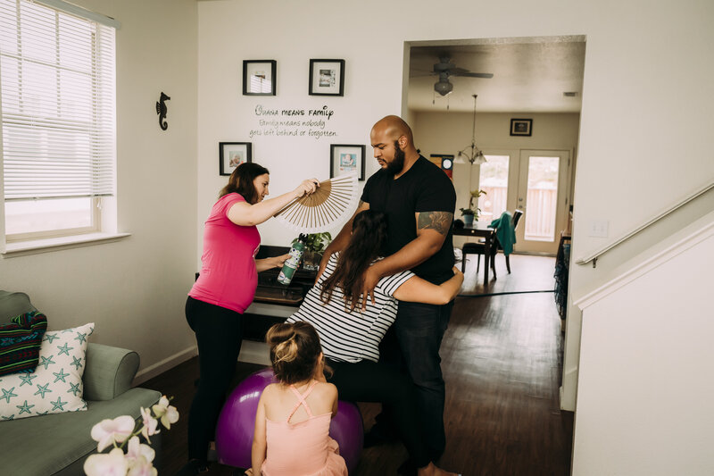 Midwife supporting laboring mother at home with partner and child present, under a sign that reads “ʻOhana means family”