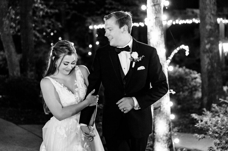 A monochrome image of an elegant bride and groom during their first dance as he wears a full traditional tuxedo and her diamond earrings sparkle in the light from the lanterns overhead.