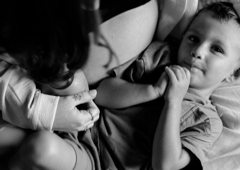 A young child lying on a pregnant woman's belly, holding her hand and looking up with a relaxed expression.