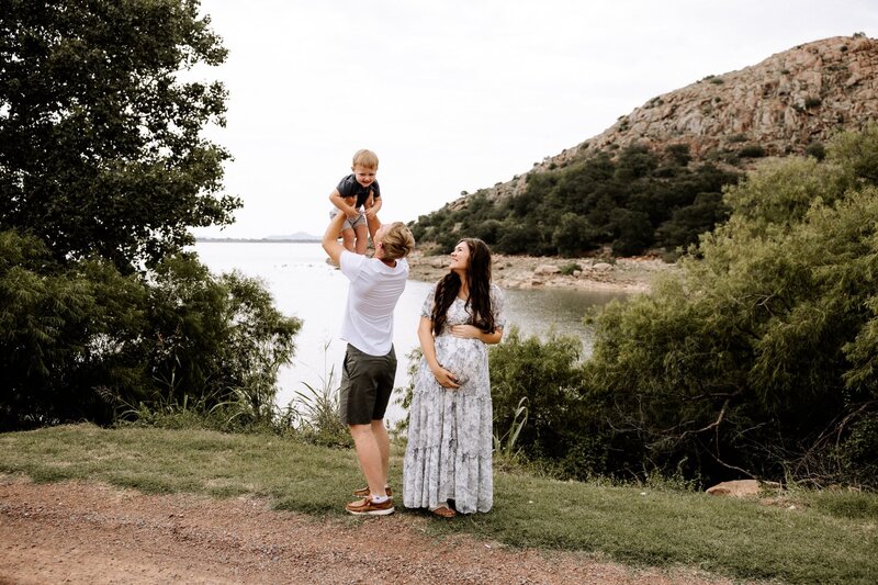 Maternity couple with a lake mountain backdrop, lake luggart in oklahoma. Father holding son above his head playing with him while mama is holding her pregnant belly.
