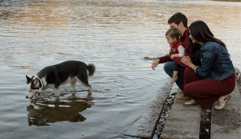 A husband and wife hold their baby and watch their dog play in the nearby lake.