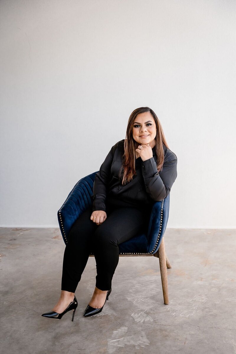 Female with long brown hair sitting in a comfortable blue chair in the middle of a plain room wearing black heels, pants and long shirt