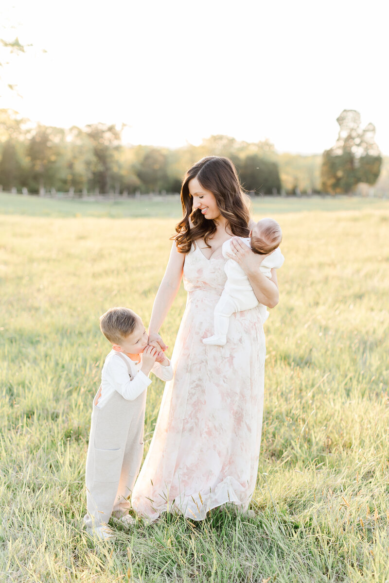 A Washington DC Photographer photo of a mother holding her toddler's hand and carrying her newborn baby outside in the Spring grass