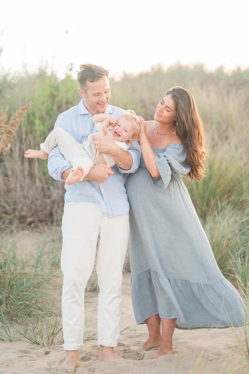 family of three on the beach for maternity photos. mom is wearing a blue flowy dress