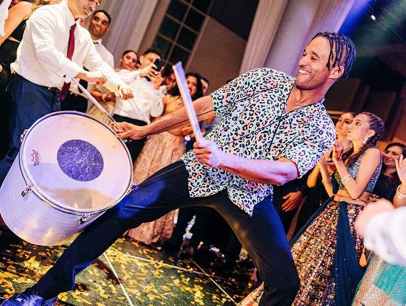 Percussionist with braided hair hyping up the dance floor while playing a large dhol-style drum surrounded by excited guests.