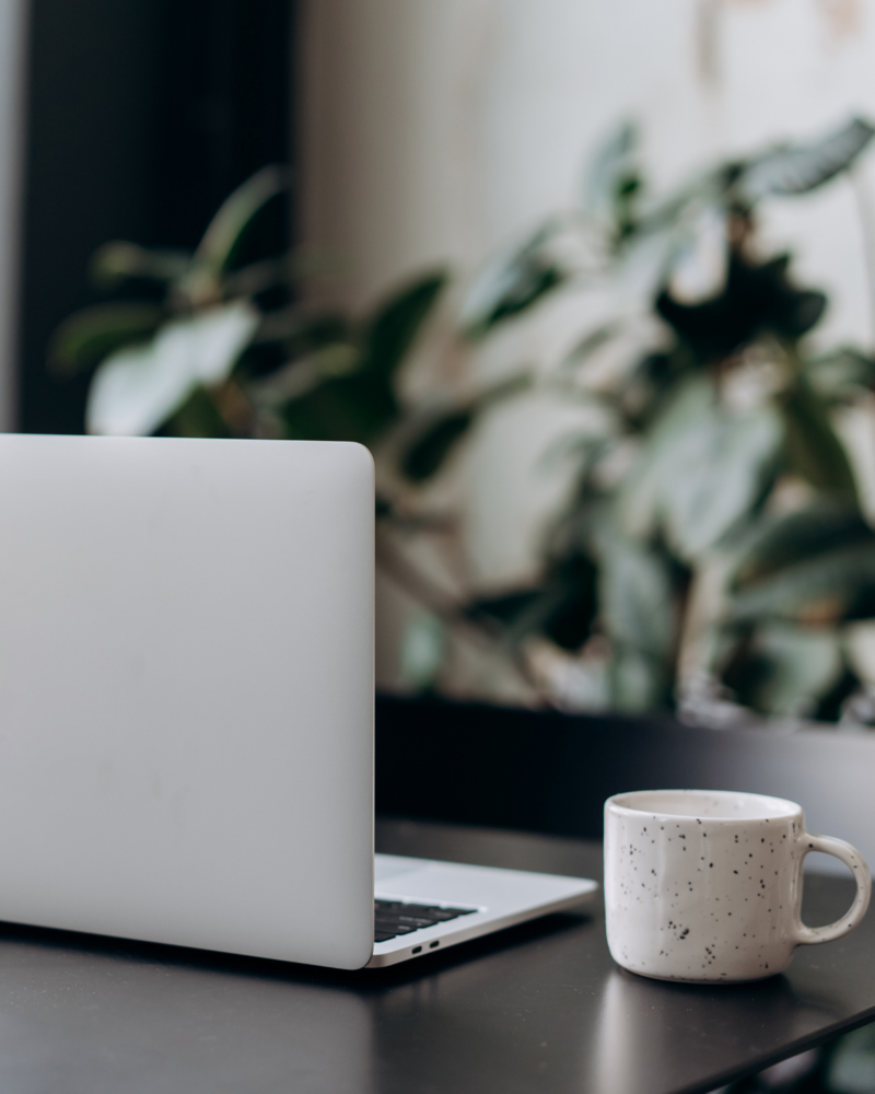 A sleek laptop and a ceramic mug on a black desk, with a blurred backdrop of indoor plants, symbolising a calm and professional telehealth environment.