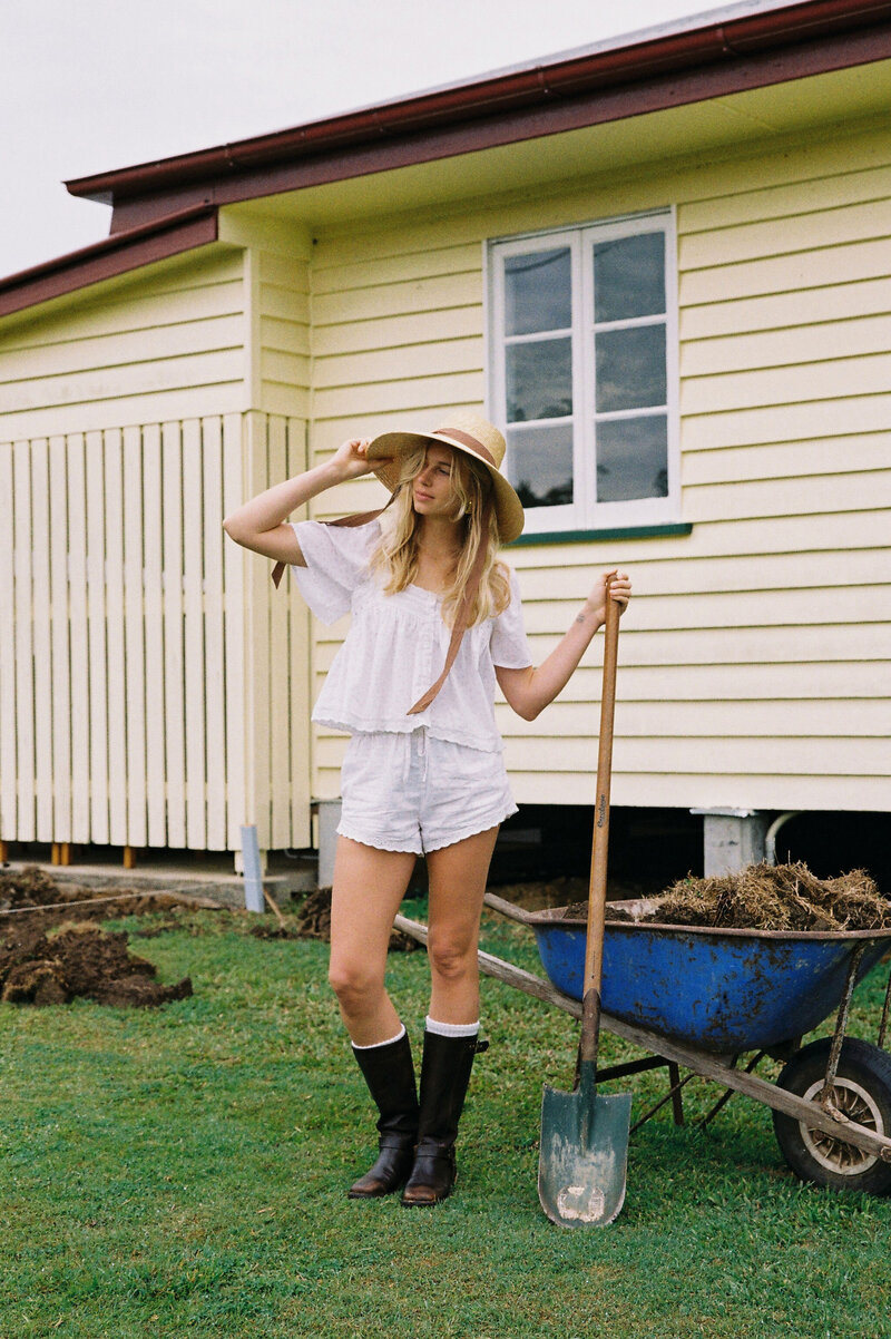 Model posing with wheelbarrow and shovel in white cotton set, farm hat and gum boots