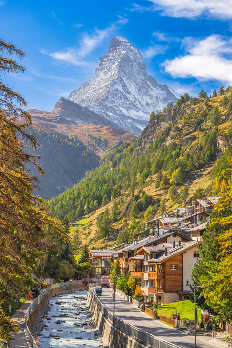 Scenic view of a Swiss village with wooden houses along a river, framed by green trees. The iconic Matterhorn mountain rises majestically in the background under a blue sky with scattered clouds.