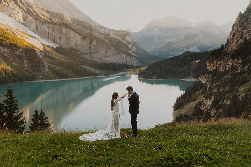 An elopement on Lake Oeschinen in Switzerland in the mountains. Man kisses his bride's hand. You are standing at the viewpoint at Oeschinensee.