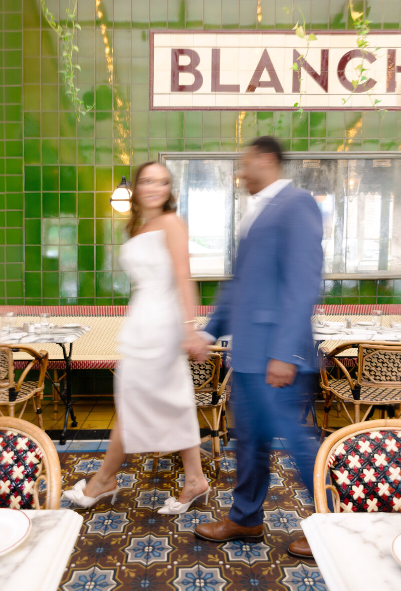 A couple holds hands walking inside a bright café with green tiled walls and a sign above. Both are in motion, creating a blur effect. The woman wears a white dress, and the man is in a blue suit. French restaurant chairs and tables fill the space.