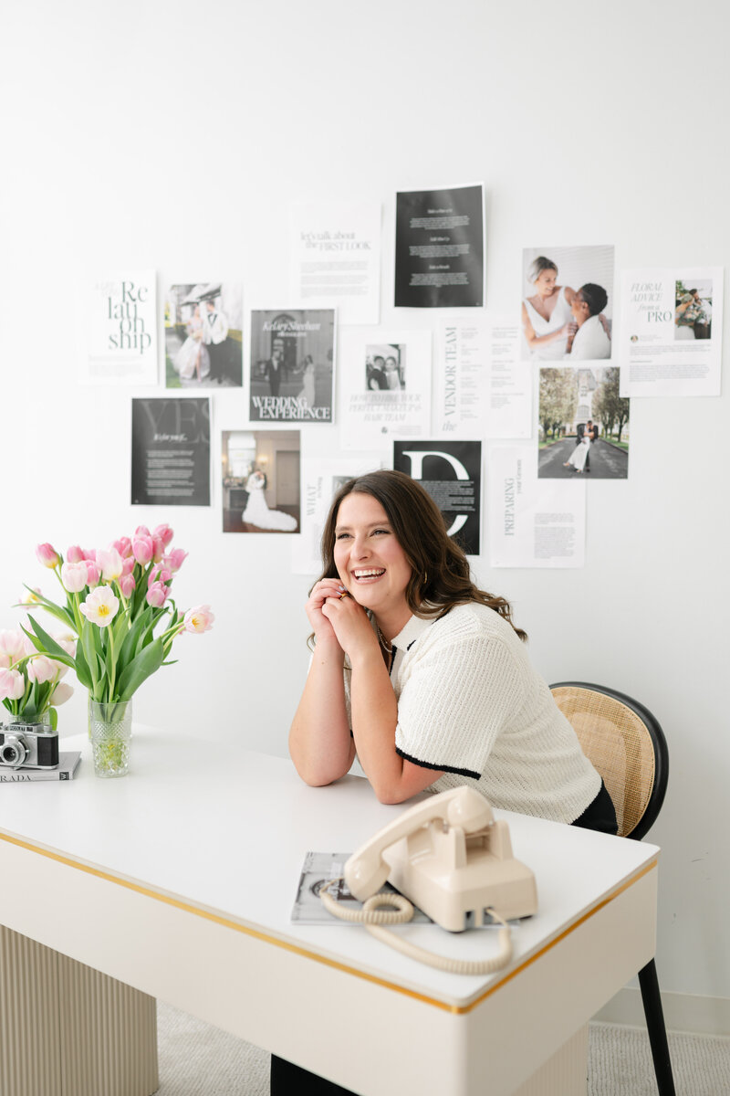 Rhode Island Wedding Photographer | Kelsey Sheehan Photography Timeless Rhode Island Weddings | Kelsey smiling, leaning on a white desk with an old-fashioned phone. Tulips in a vase, and a collage of photos and quotes on the wall add an artistic touch.
