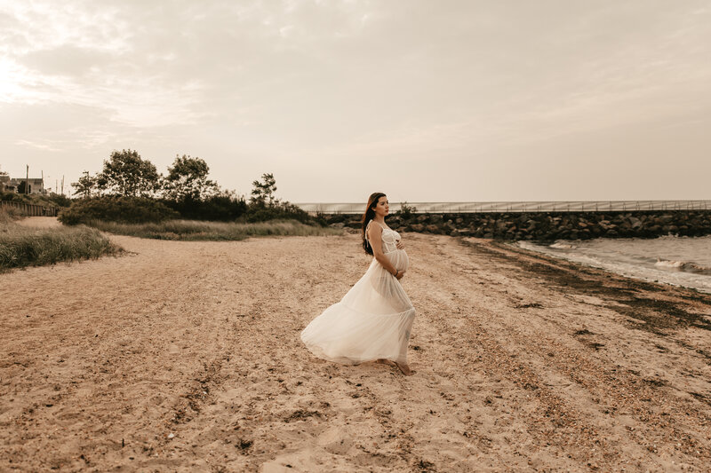 Pregnant woman in a sheer white dress posing on a sandy beach during a sunset maternity session in Old Bridge, NJ, captured by Burkat Photography.