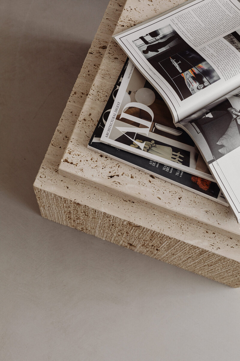 A stack of travertine furniture topped with opened and stacked Vogue magazines. The clean, neutral-toned stone contrasts with the crisp magazine pages.
