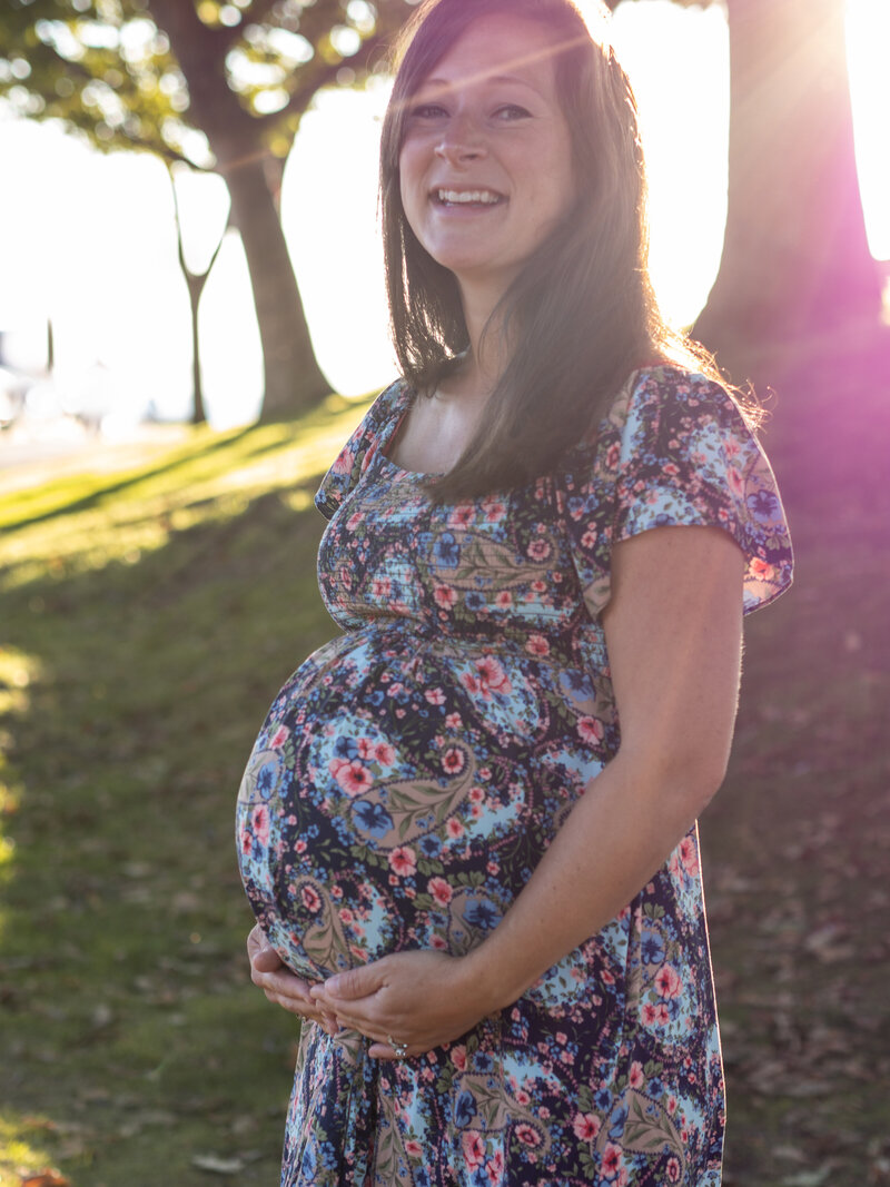 A pregnant woman is standing in Edmonds, WA smiling as she holds her protruding belly.