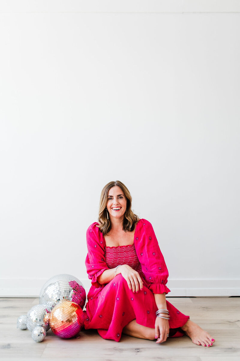 female photographer wearing pink pants and white top sitting next to a disco ball