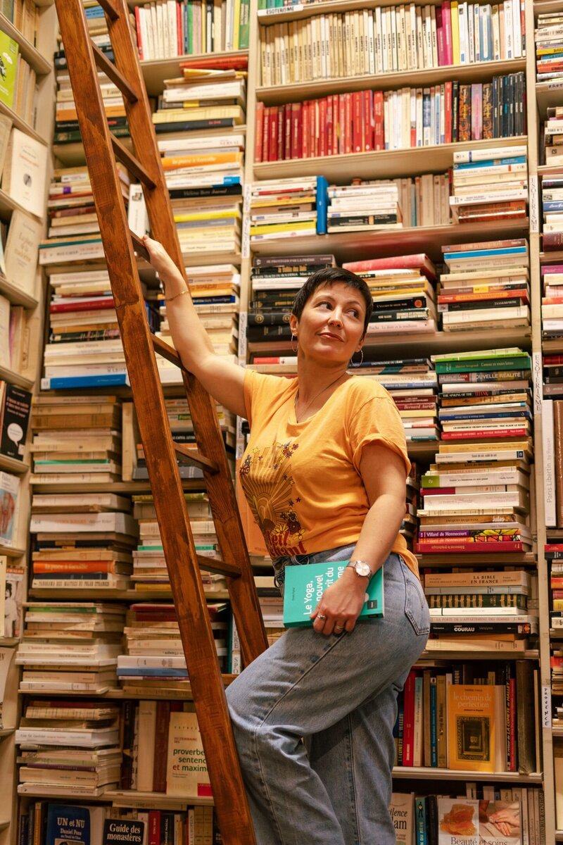 Leslie est sur une échelle de libraire avec un livre à la main. En arrière-plan, les étagères pleines de livres de la librairie.