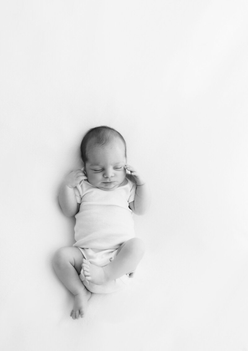Black and white photo of a newborn baby lying on their back on a soft, light-colored surface. The baby is wearing a simple onesie and has a peaceful expression with eyes closed, tiny legs slightly bent, and hands resting beside their head. Captured by a talented St. Louis photographer.