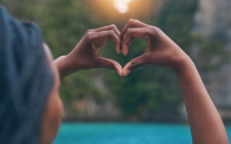 woman holding her hands in the shape of a heart near a body of water surrounded by green foliage