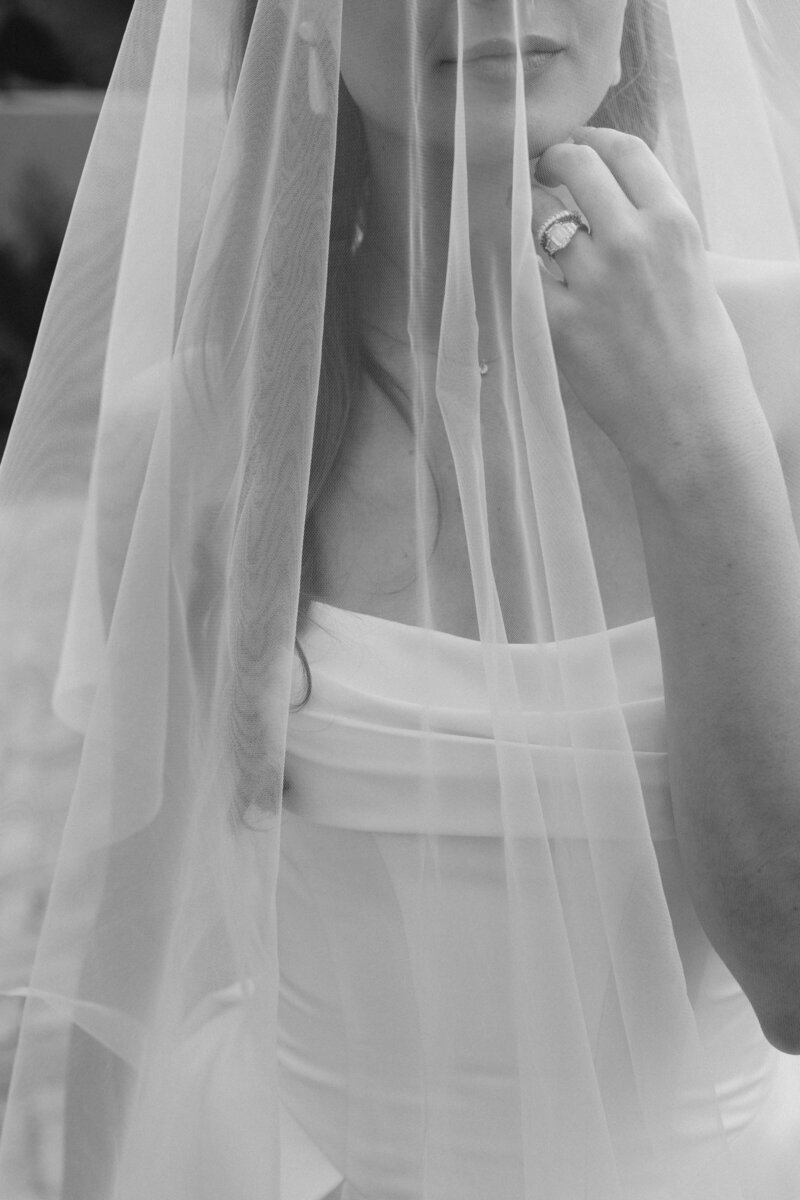 Black and white photo of bride holding her chin underneath her veil with her rings showing