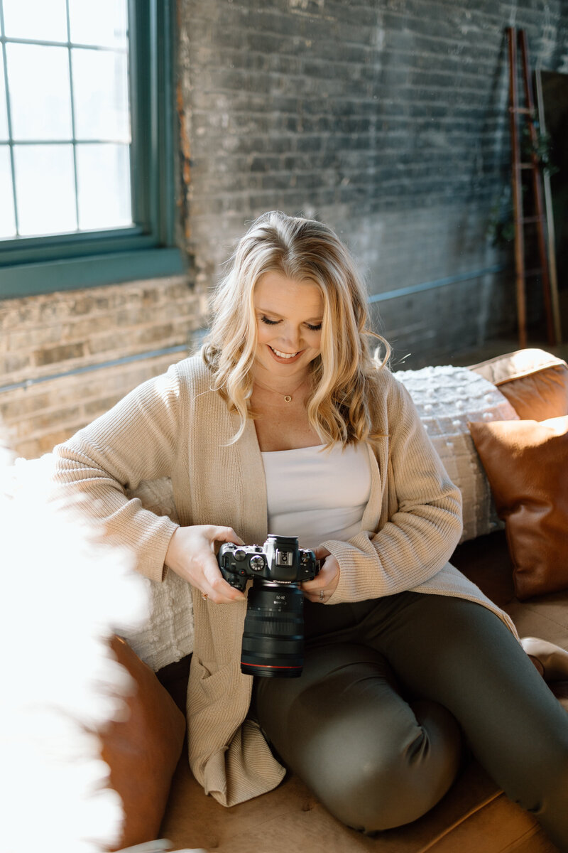 photographer smiles down at her camera while seated on a tan couch in an industrial setting