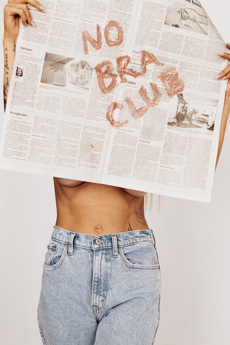 A shirtless girl with a newspaper covering her that says no bra club. 