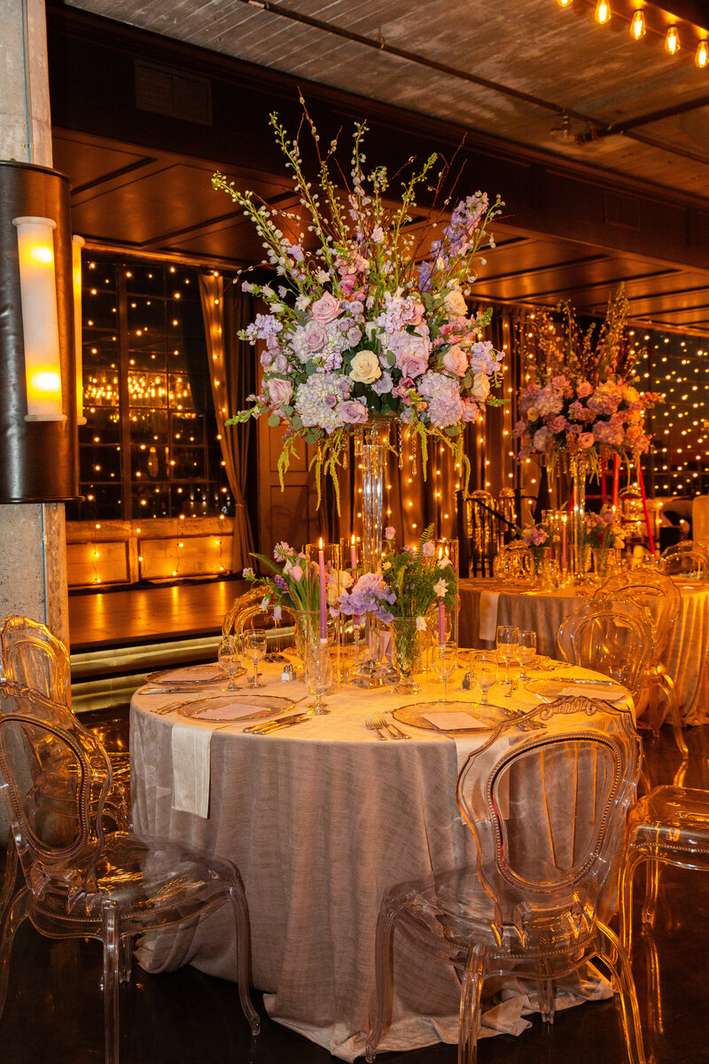 Indoor reception setting with round tables and ghost chairs, accented by tall floral centerpieces and twinkle lights.
