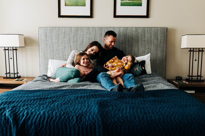 a mother, father, and two young girls snuggle on a bed during a family portrait session in houston texas