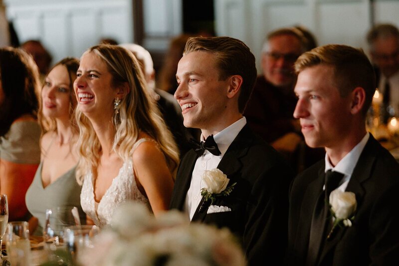 A newlywed couple laughing as they sit at a table with their wedding party 