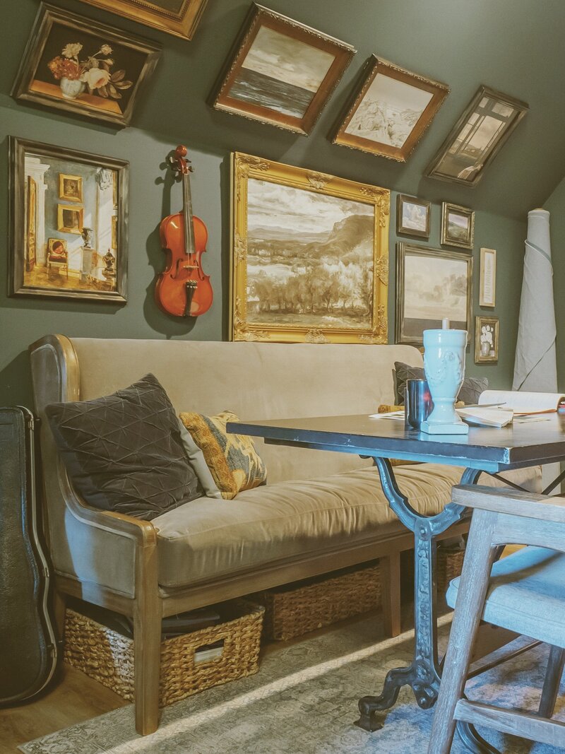 Gallery wall with artwork of nature scenes, home scenes and violin on the wall with slanted ceiling, a sofa bench with pillows and a wrought iron table with chairs and office items strewn on the table