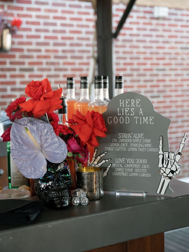 black skull vase with rave jeweled "makeup" and red roses, iridescent anthurium, and hot pink carnation arrangement sitting on a bar next to a "Here Lies a Good Time" signature cocktail tombstone bar sign