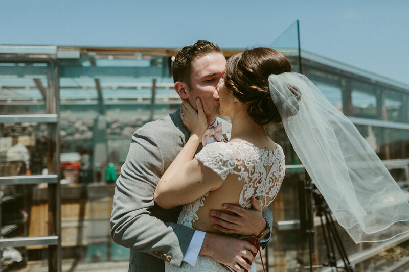 A bride and groom share a passionate kiss on a rooftop venue, with a modern glass structure reflecting the sunlight.