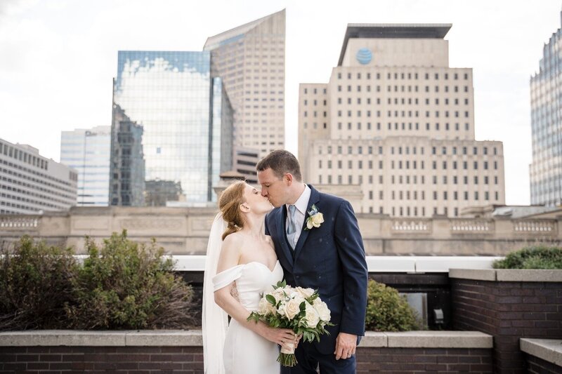 Bride and groom share a romantic kiss on the rooftop at JPS Events in downtown Indianapolis, with a city skyline and modern high-rise buildings in the background. The bride holds a bouquet of white roses while wearing an off-the-shoulder gown.