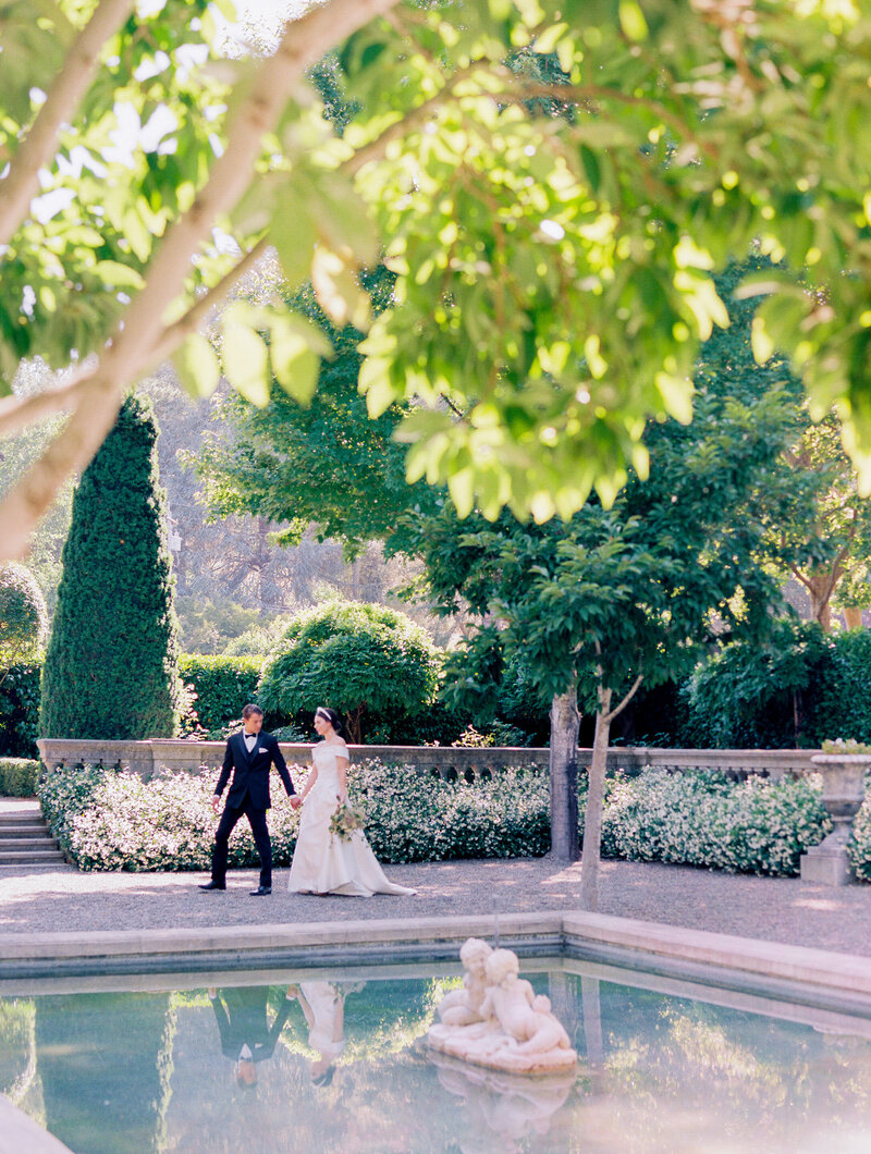 bride and groom walking frmo far away with trees framing next to fountain. bride has pearl headband