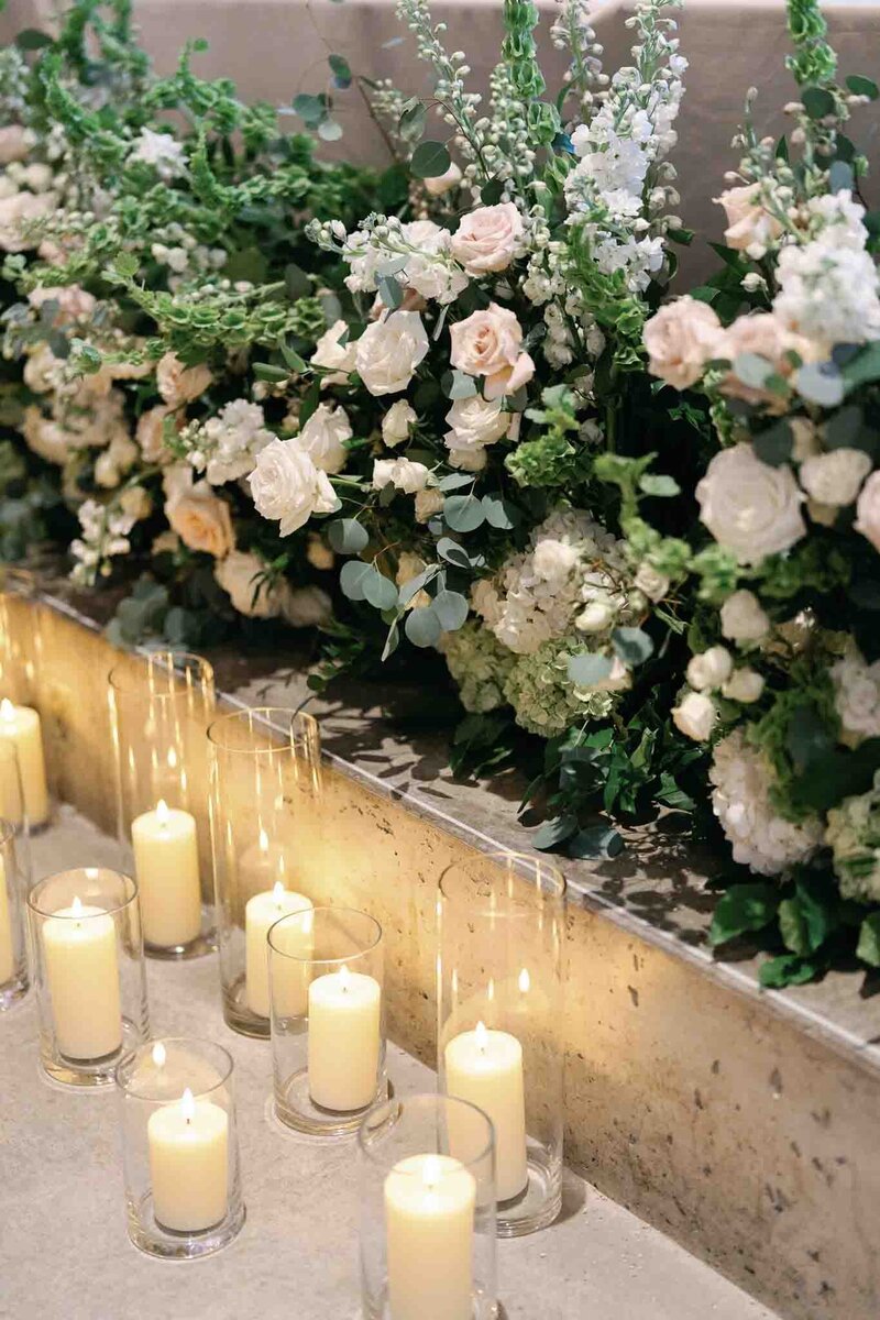photo of greenery with white and blush florals sitting on a concrete ledge with tall glass vases with cream colored candles in them below the florals