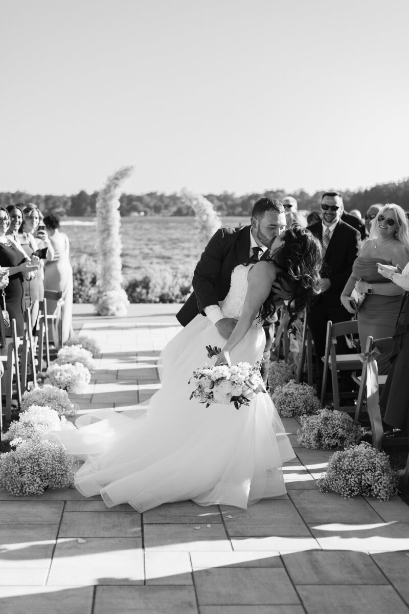 Rhode Island Wedding Photographer | Kelsey Sheehan Photography Timeless Rhode Island Weddings | A newlywed couple shares a joyful kiss on an outdoor aisle, surrounded by guests. The bride holds a bouquet and wears a flowing gown. The scene exudes happiness.
