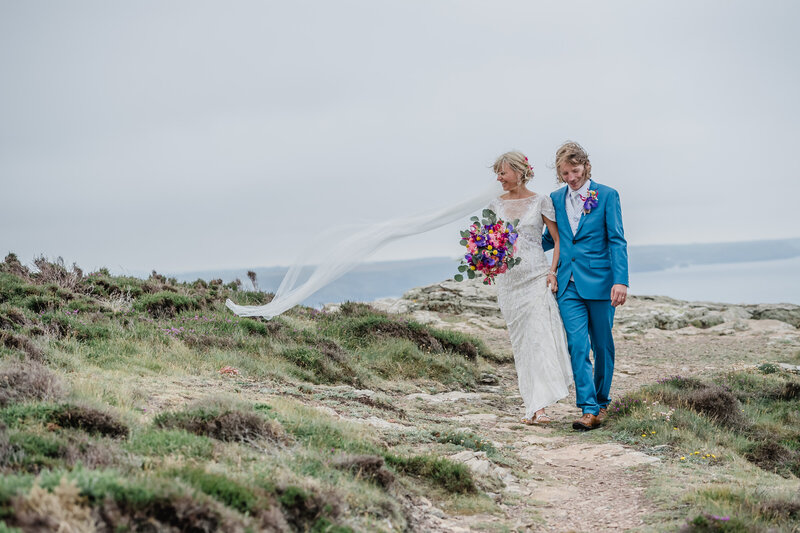 A newly married couple walking along the cliff tops in Cornwall. The bride is carrying a bouquet of purple orchids, yellow crespedia and pink roses