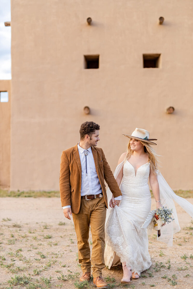 Couple just married in western style wedding dress. They are walking and looking each other. In the background the Fort of Fort Lupton.