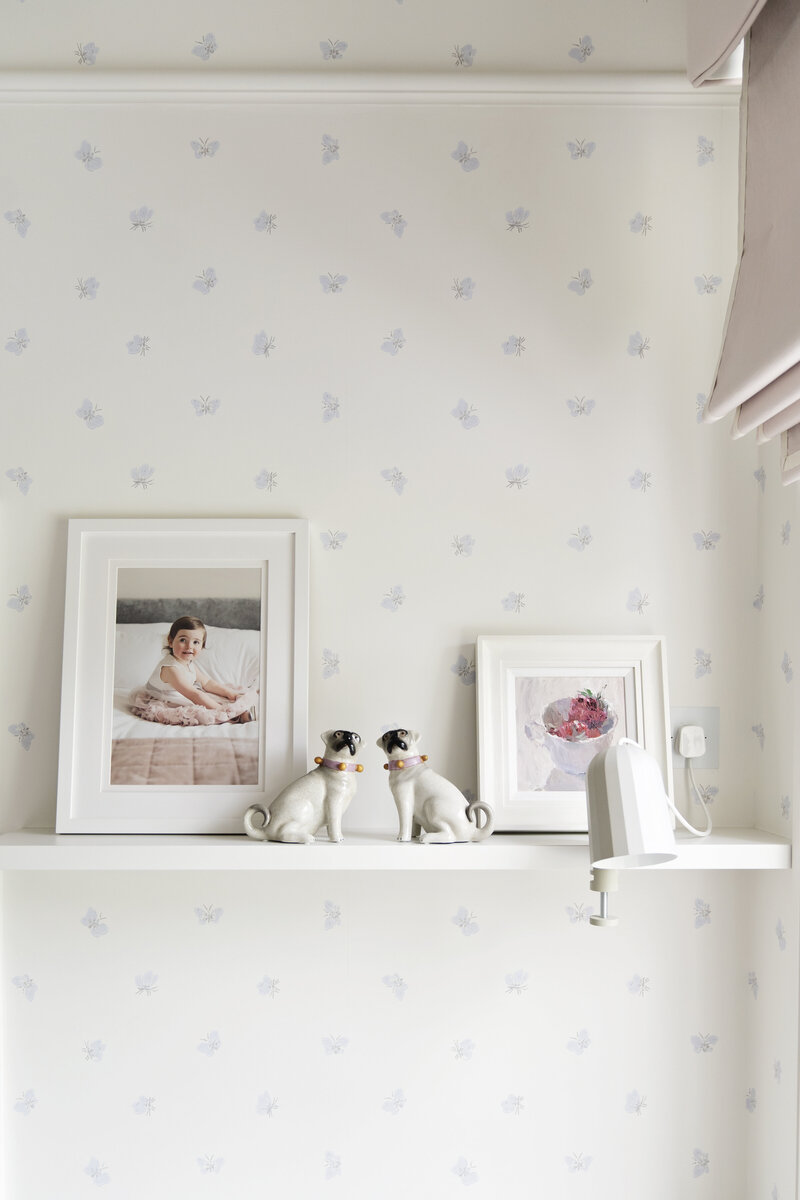 A framed photo of a little girl by Jess Morgan Photography sits on a shelf along with some china pugs and a small painting of fruit in a bowl.
