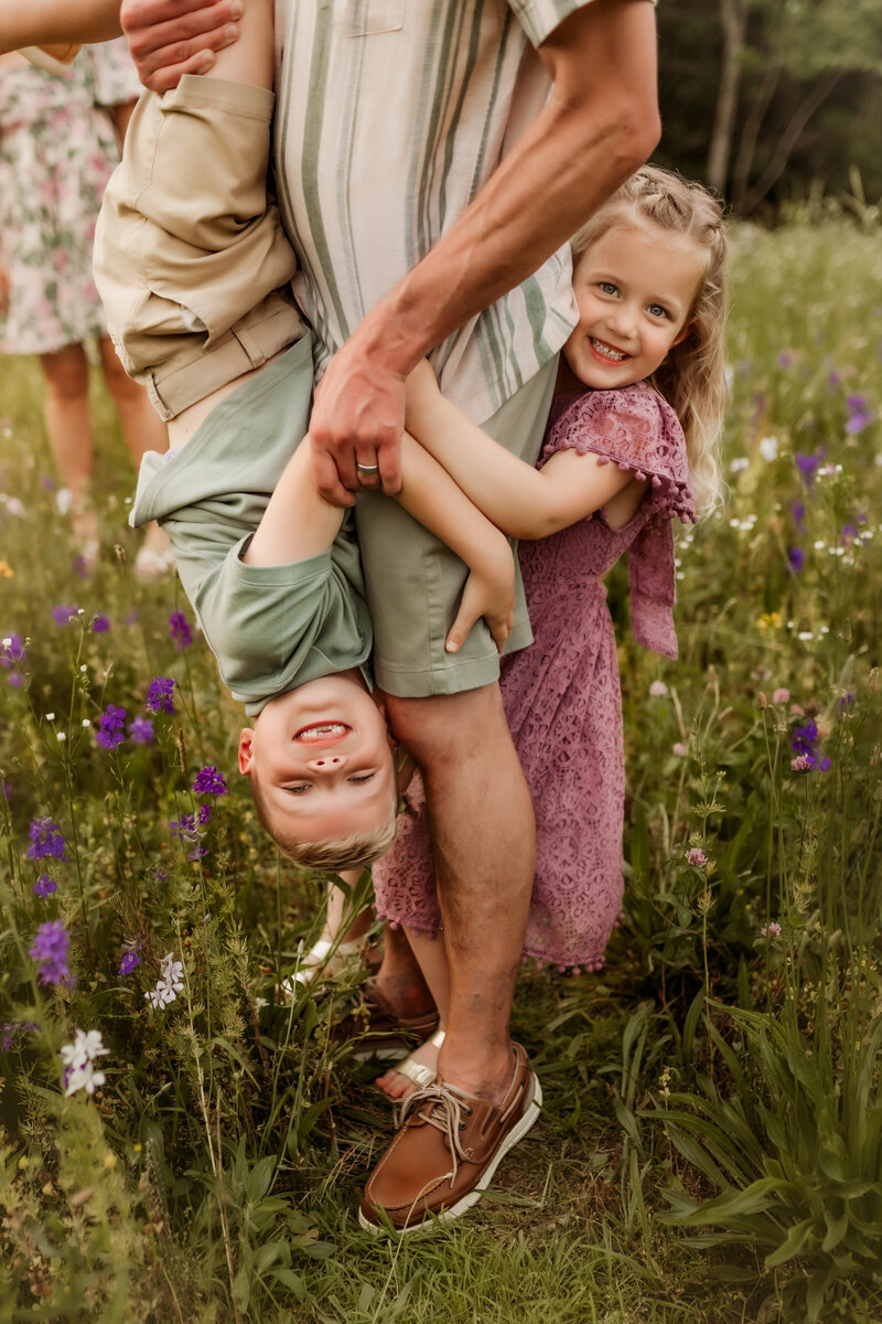 Little boy hangs upside down as little sister hugs their Dad from behind while Shawnee Photographers captured their smiles